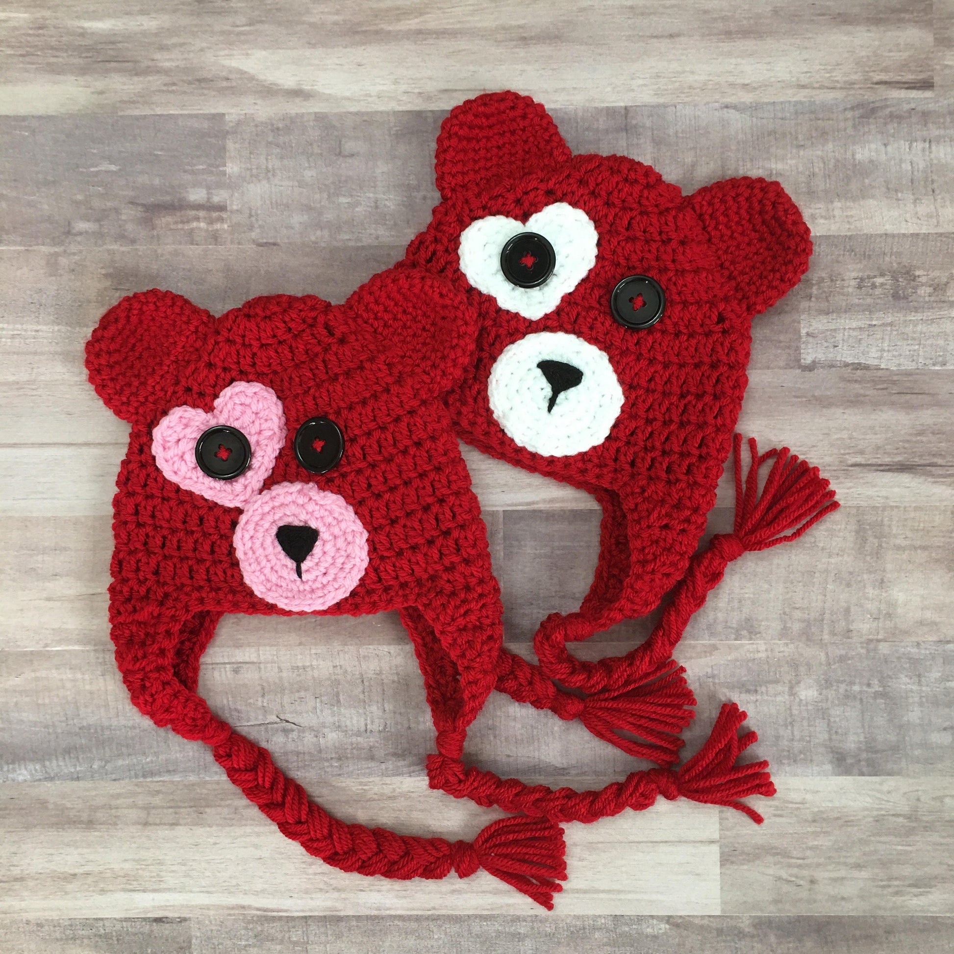 Two red bear hats. One with a white heart around the eye and muzzle. One with a pink heart around the eye and muzzle